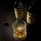Two bottles of Jameson whiskey with a glass filled with whiskey on a dark surface.