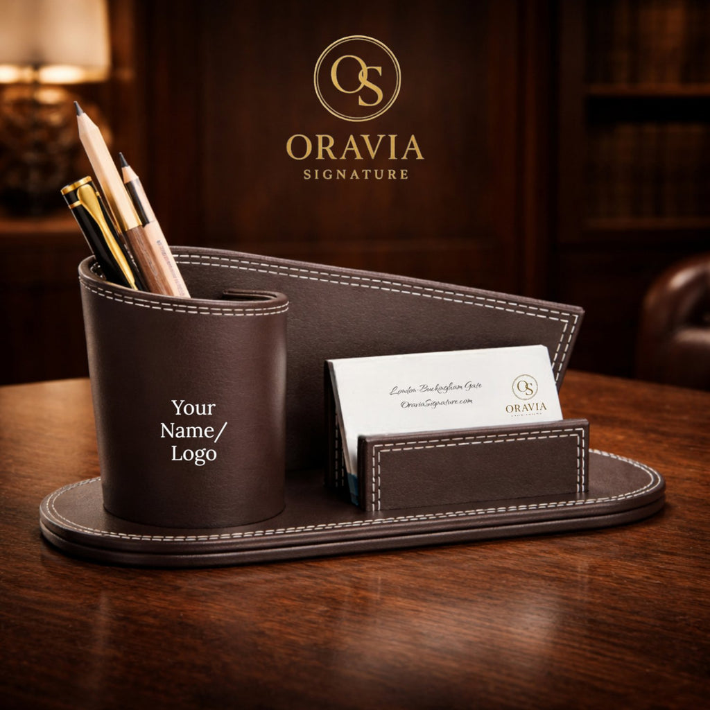 Brown leather desk organizer with pens and a card on a wooden surface, featuring the Oravia Signature logo.