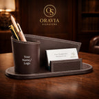 Brown leather desk organizer with pens and a card on a wooden surface, featuring the Oravia Signature logo.
