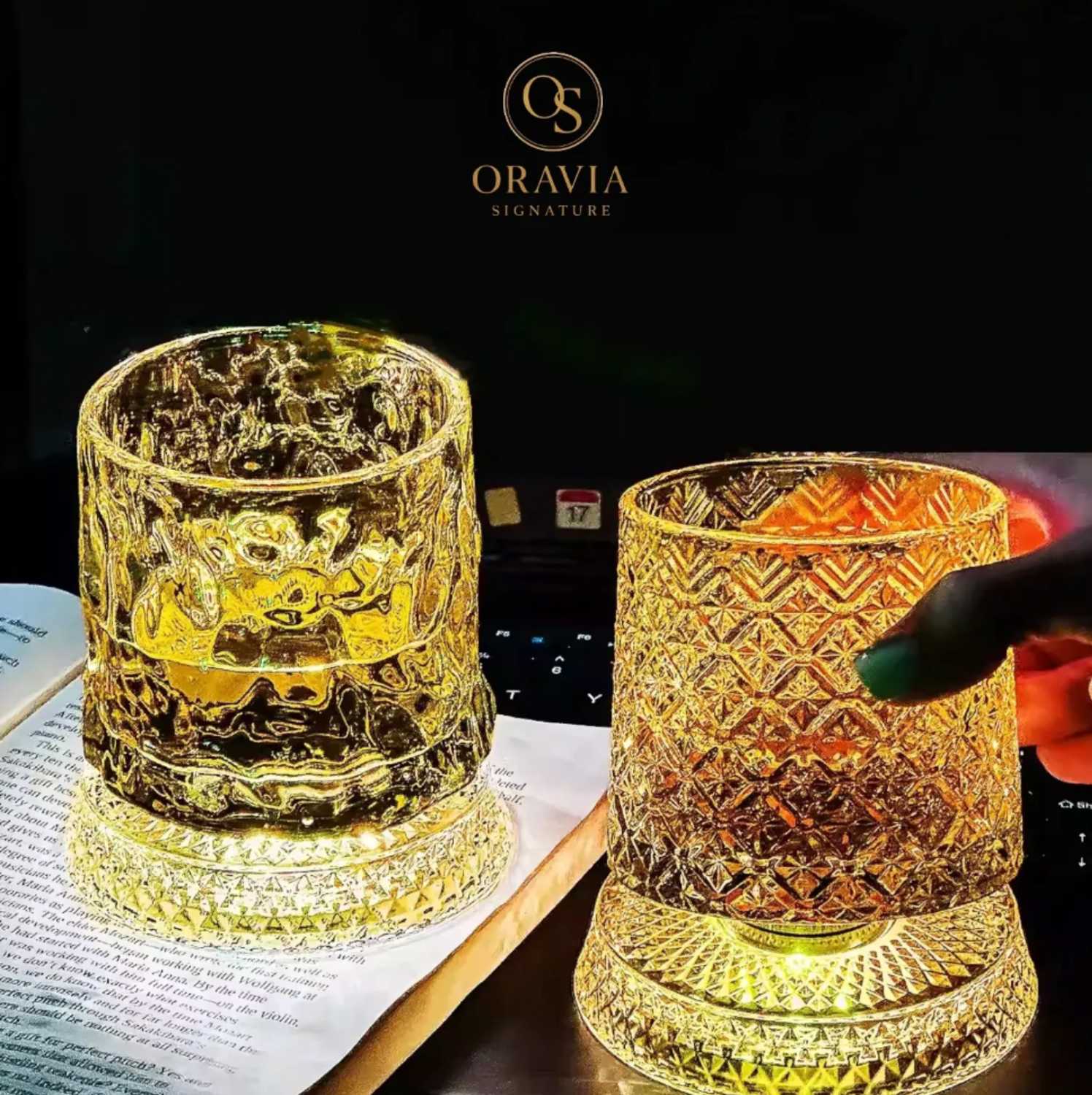 Two illuminated glass candle holders with intricate designs on a dark background, featuring the Oravia brand.