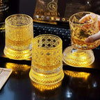 Three ornate glass tumblers with a hand holding one, on a reflective surface with a blurred background.