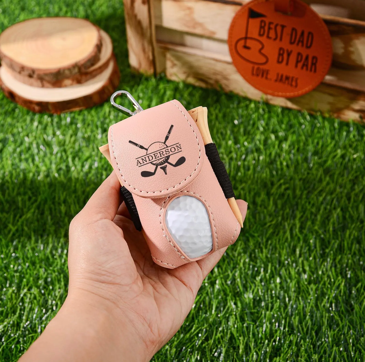 Hand holding a pink golf ball and club head cover with a golf-themed design on grass.