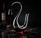 Glass wine decanter shaped like a swan with red wine on a dark surface, with a bottle and glass in the background.