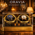 Two watches in a gold display case with 'ORAVIA' branding in an elegant room setting.